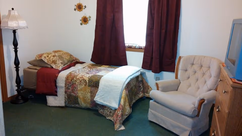 A small bedroom with a single bed covered in a patchwork quilt and pillows, a tall floor lamp with a white lampshade, a beige upholstered armchair, a wooden dresser with a blue TV on top, and a window with dark red curtains.