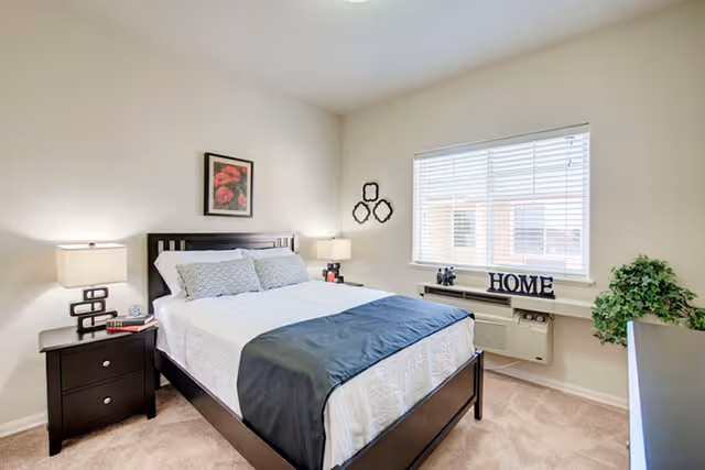 A bright bedroom with a neatly made bed featuring white and gray bedding and a dark wooden headboard. There are two matching dark wooden nightstands with modern lamps on each side of the bed. A framed floral picture hangs above the headboard. A window with white blinds lets in natural light, and a shelf below the window holds decorative items including the word 'HOME'. A green potted plant is placed near the window, and the room has beige carpet flooring.