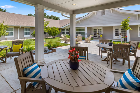 Covered courtyard patio with round wooden tables, chairs with striped cushions, potted flowers, and walkways in front of a single-story building.
