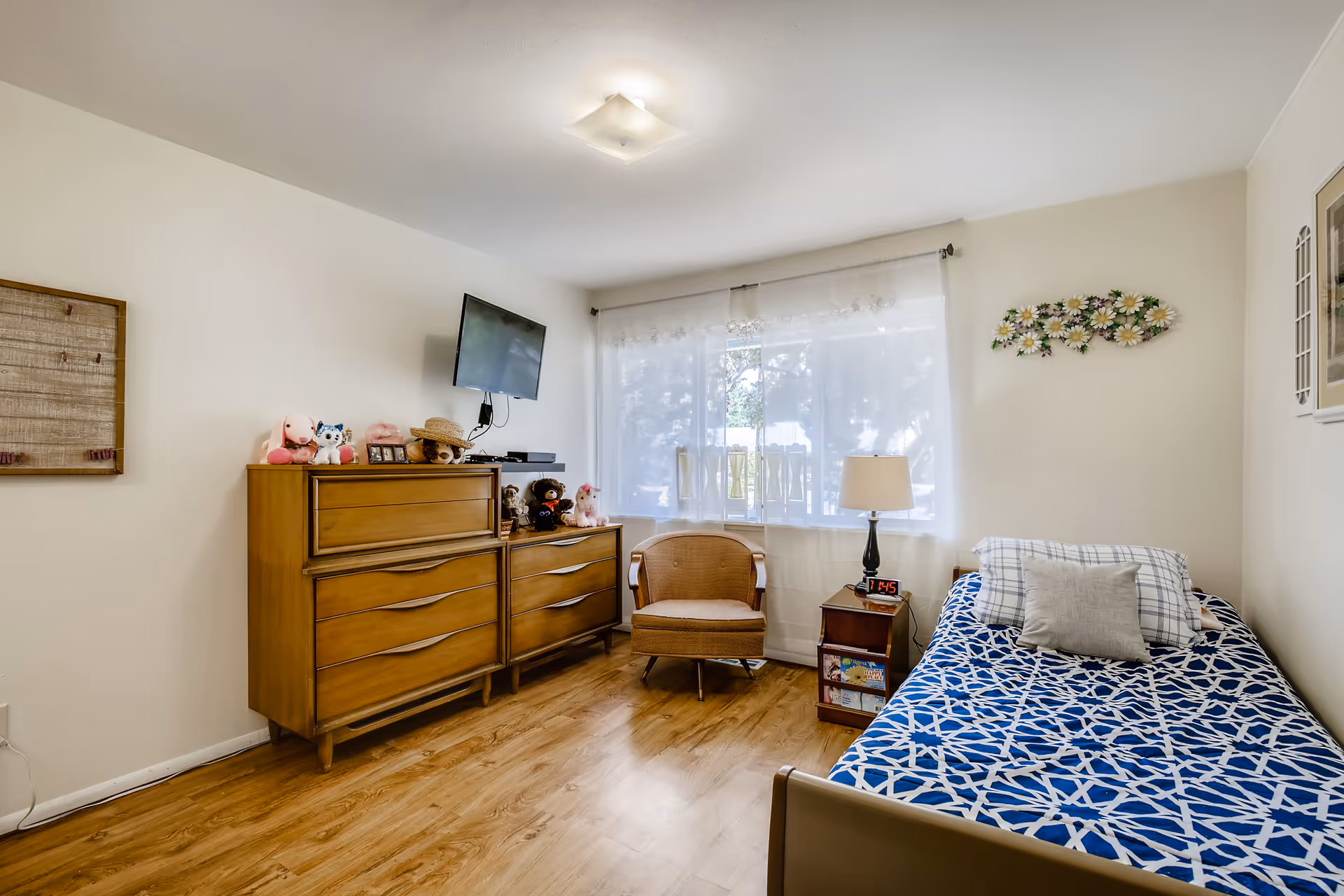 A cozy bedroom with a single bed covered in a blue and white patterned bedspread, a wooden nightstand with a lamp and digital clock, a wicker chair, and two wooden dressers topped with stuffed animals and a mounted flat-screen TV. The room has light-colored walls, a large window with sheer white curtains, and wooden flooring.