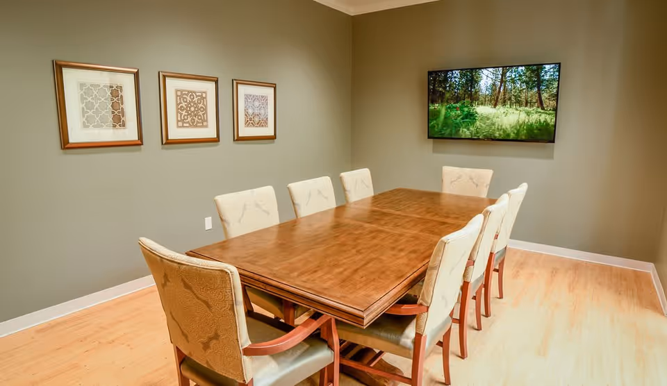 A dining room with a long wooden table surrounded by eight upholstered chairs. On the wall above the table are three framed decorative artworks, and a flat-screen TV displaying a nature scene is mounted on the adjacent wall. The room has light wood flooring and beige walls.