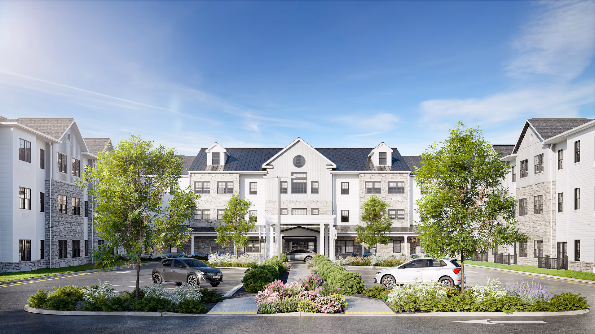 Front exterior of a three-story senior living building with a landscaped circular drive, trees, and parked cars under a blue sky.