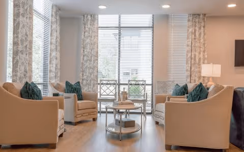 A bright and cozy living room with large windows covered by patterned curtains, four beige armchairs with green throw pillows arranged around a round glass coffee table, and a floor lamp in the corner.