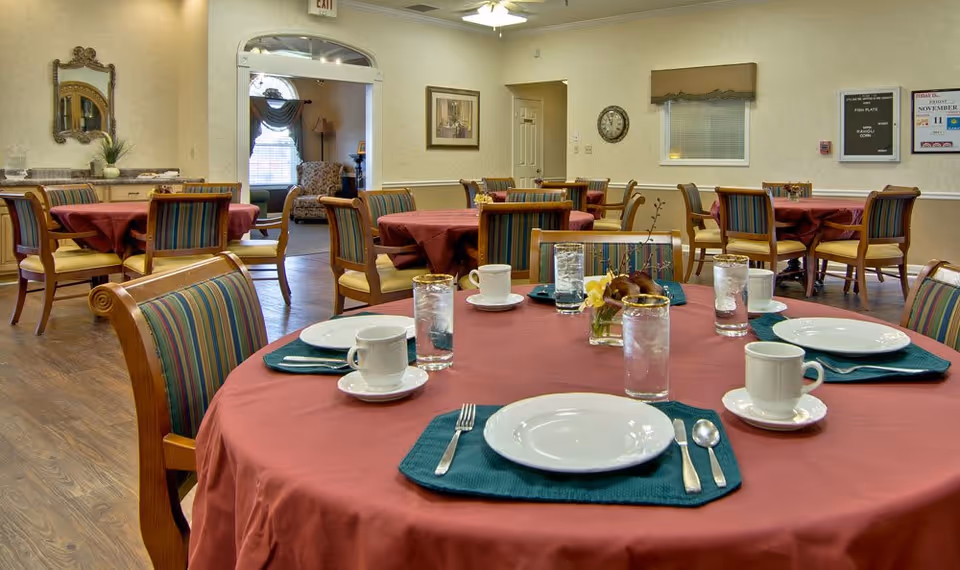 Dining room with round tables covered in red tablecloths, each set with white plates, cups, silverware, and glasses of water. Chairs with striped upholstery surround the tables. The room has beige walls, a wooden floor, and a doorway leading to a sitting area with armchairs and a window.