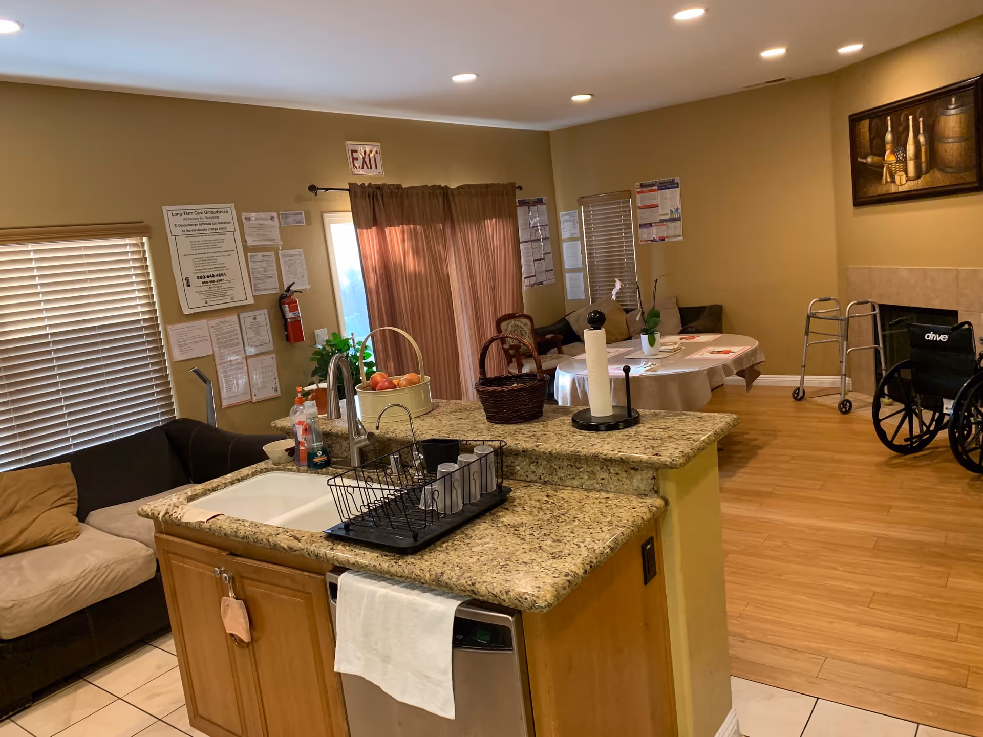 Interior view of a senior living facility common area featuring a kitchen island with a sink, dish rack, and paper towel holder. In the background, there is a round table with chairs, a couch, a wheelchair, a walker, and various notices and decorations on the walls. The room has wooden flooring and beige walls with a painting above the fireplace.