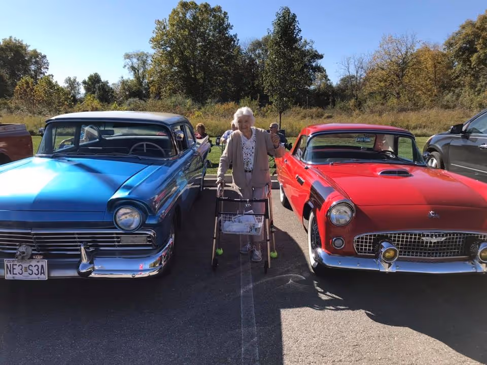 An elderly woman using a walker stands between a blue and a red classic car in a sunny parking lot with trees behind.