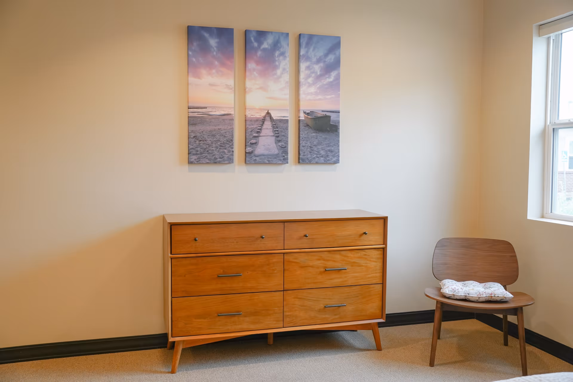 Wooden dresser and chair beneath a three-panel beach artwork next to a window in a simple room.