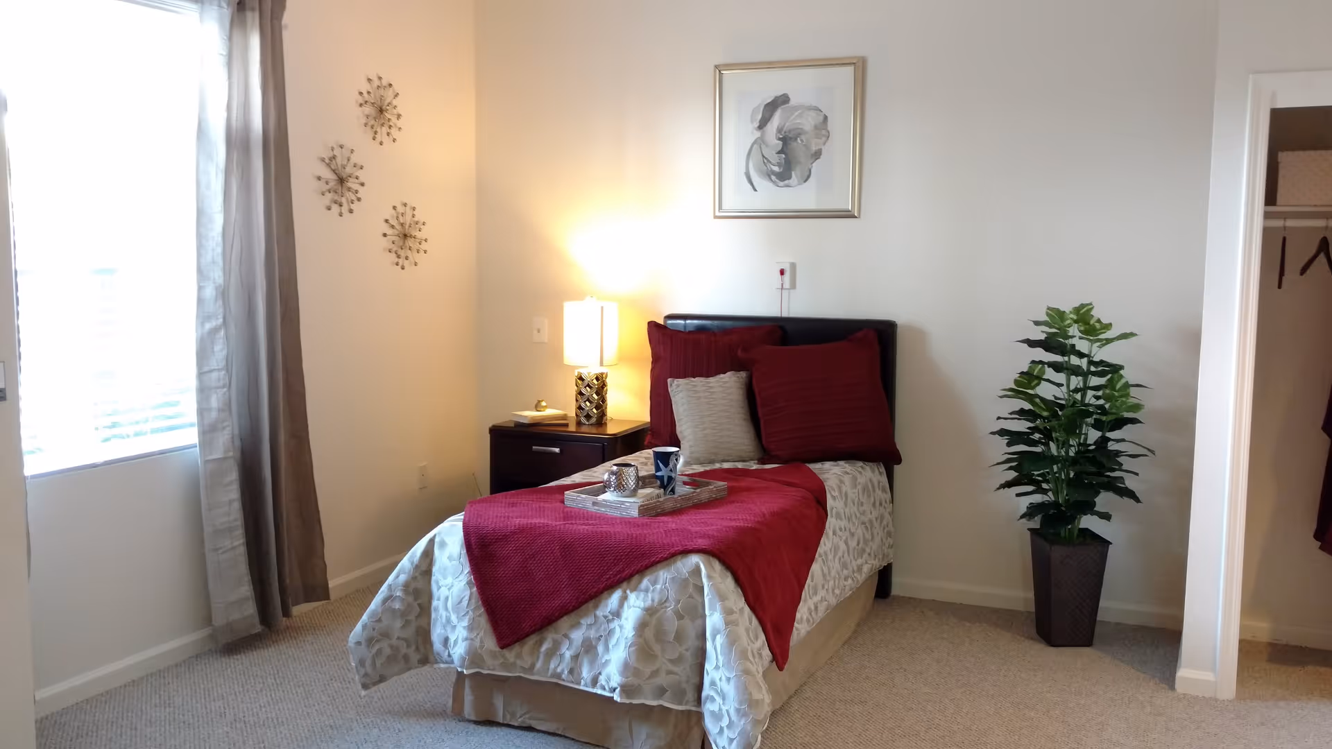 A cozy bedroom with a single bed dressed in white and red bedding, two red pillows and one beige pillow. A wooden nightstand with a lamp and a small decorative item sits next to the bed. There is a framed abstract painting on the wall above the bed. A green potted plant is placed on the floor near an open closet. Light beige carpet covers the floor and a window with beige curtains allows natural light into the room.