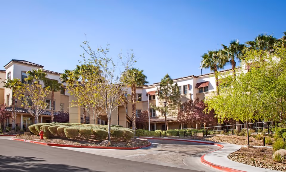 Exterior view of a multi-story senior living facility with beige walls, red-tiled roofs, and multiple windows. The building is surrounded by landscaped greenery including trimmed bushes, palm trees, and other trees. A curved driveway with red curbs leads to the entrance under a clear blue sky.
