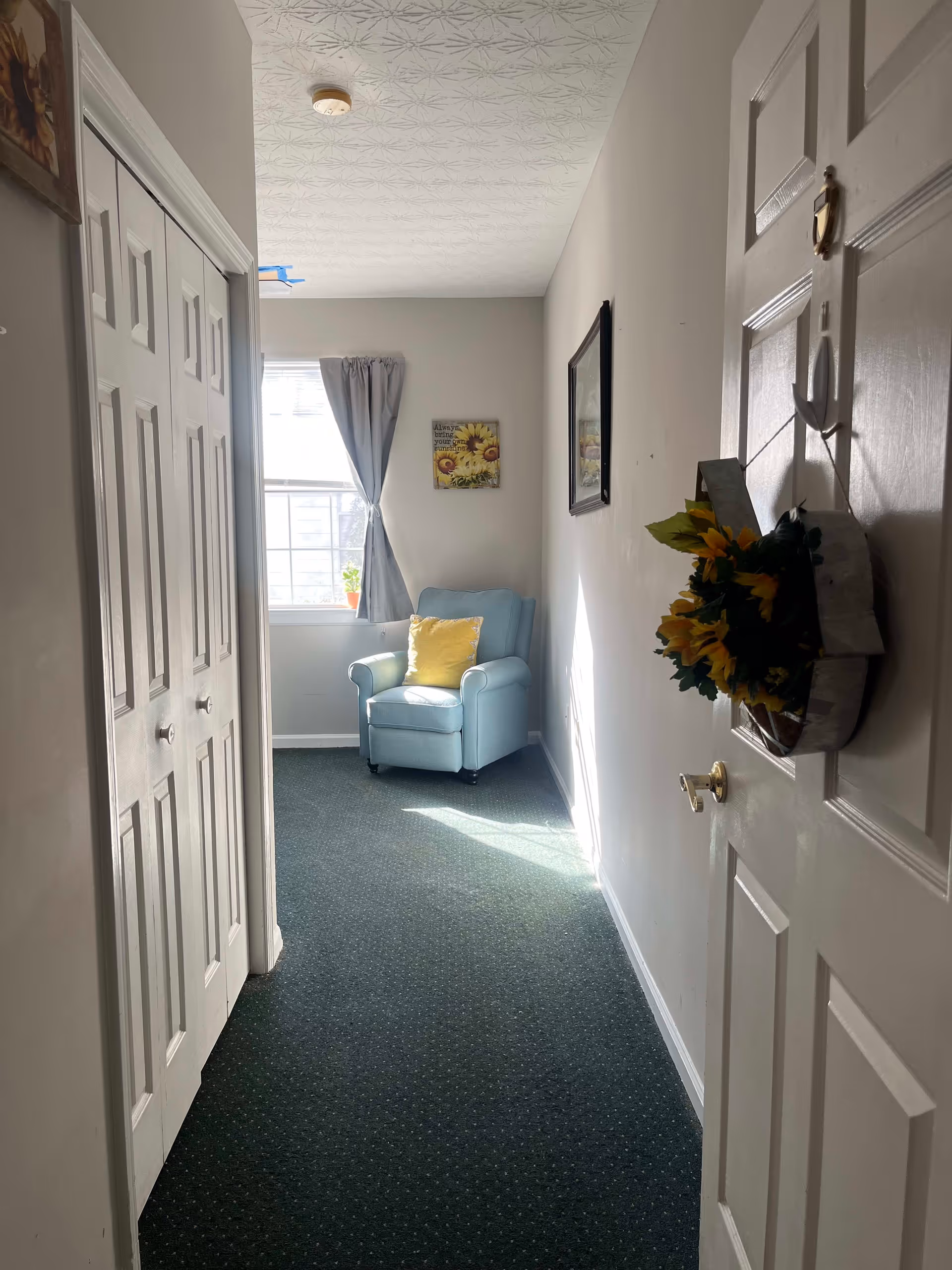 A hallway with a closed white door on the right decorated with a sunflower wreath. On the left side, there are white closet doors. At the end of the hallway, there is a window with gray curtains and a light blue armchair with a yellow pillow placed in front of it. The walls are light-colored and the floor is covered with dark green carpet.
