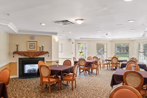 Dining room with multiple round tables and chairs, a fireplace on the left, and windows overlooking a lawn.
