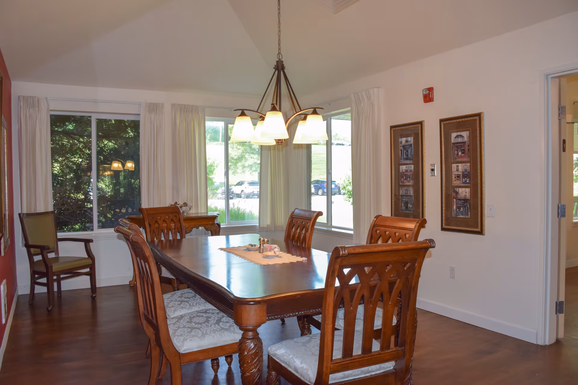 A dining room with a wooden dining table surrounded by six matching chairs with cushioned seats. A chandelier with five lights hangs above the table. Large windows with white curtains allow natural light to fill the room. There is a wooden chair with armrests against a red accent wall on the left, and two framed pictures hang on the white wall to the right. The floor is wooden, and a small table with decorative items is visible near the windows.
