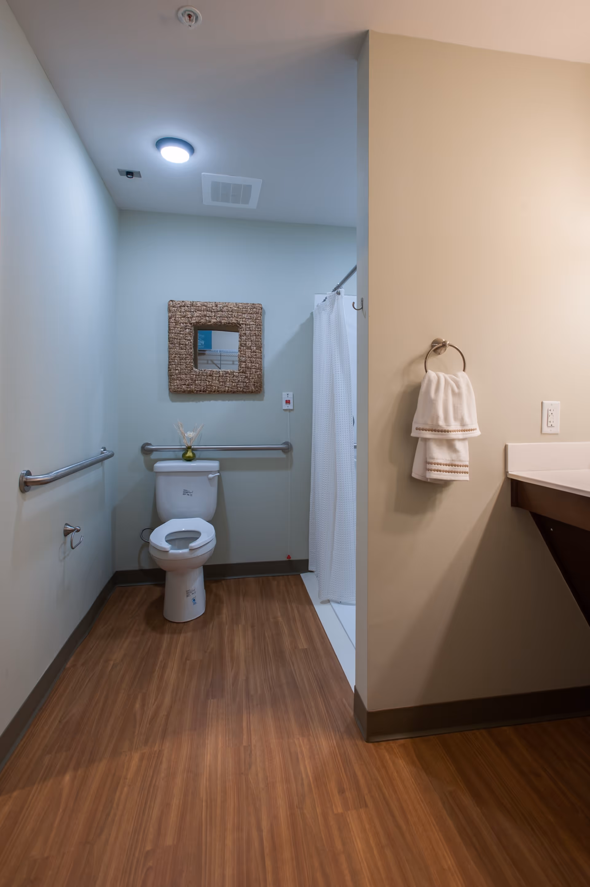 A clean and accessible bathroom with a toilet, grab bars on the walls, a small mirror above the toilet, a white shower curtain, and a towel hanging on a ring near a sink with a countertop.