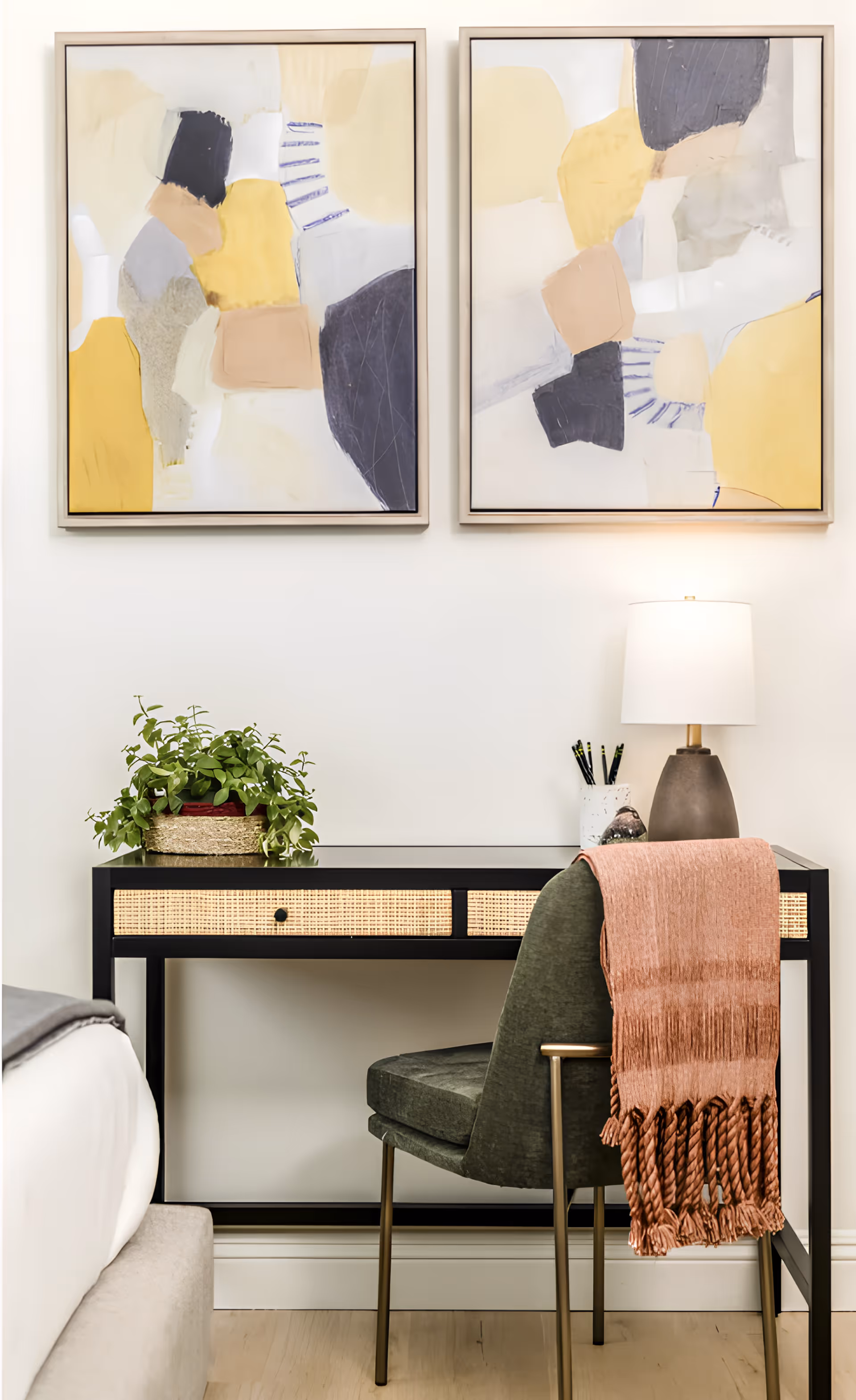 A small desk and chair with a lamp, plant and framed abstract art above, next to a bed.