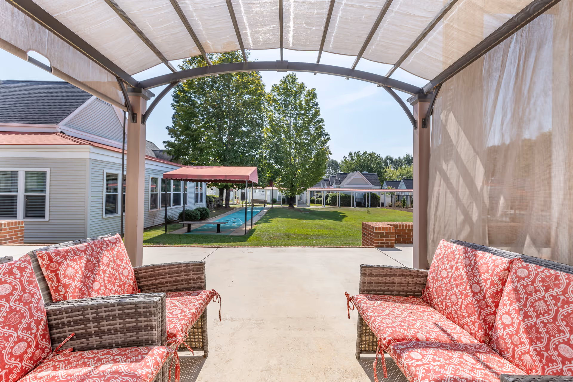 Outdoor seating area with two wicker sofas featuring red patterned cushions under a shaded canopy, overlooking a green lawn with trees and a shuffleboard court, adjacent to light-colored buildings with windows.