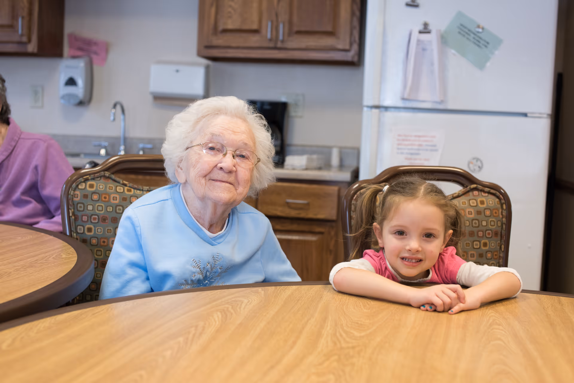 An elderly woman and a young girl sitting at a round wooden table in a communal dining area.