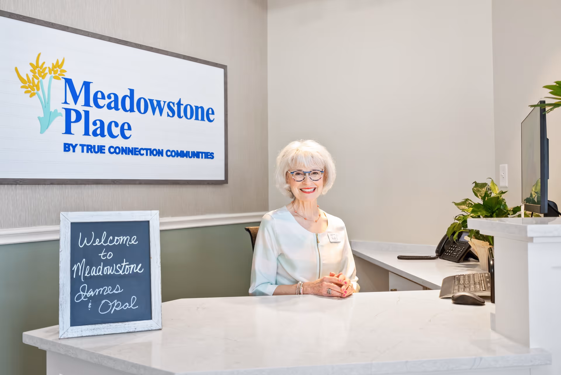 Reception area of Meadowstone Place senior living facility with a smiling elderly woman sitting behind a white marble counter. A sign on the wall reads 'Meadowstone Place by True Connection Communities' and a small chalkboard on the counter says 'Welcome to Meadowstone James & Opal'.