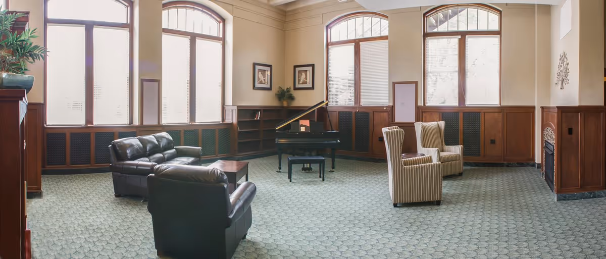 A spacious living room with large arched windows letting in natural light. The room features a black grand piano with a matching bench, two striped armchairs, a black leather sofa, and a black leather armchair arranged around a small wooden coffee table. The walls have wood paneling on the lower half and light-colored paint above, with framed artwork and a decorative plant on a built-in bookshelf. There is also a fireplace on the right side of the room.