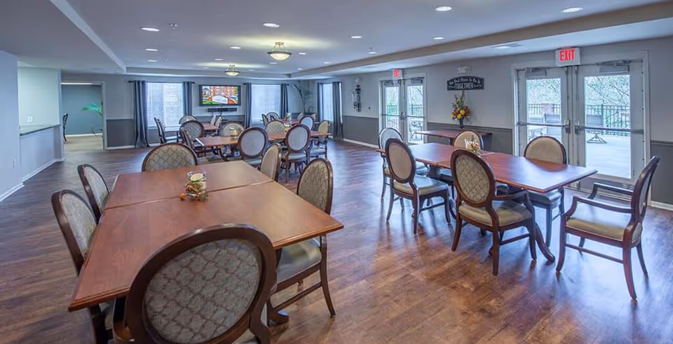 A spacious dining room with multiple wooden tables and cushioned chairs arranged neatly. The room has large windows with curtains, allowing natural light to enter. There are double glass doors leading to an outdoor area. The floor is wooden, and the walls are painted in light colors with some decorations and a TV mounted on the far wall.