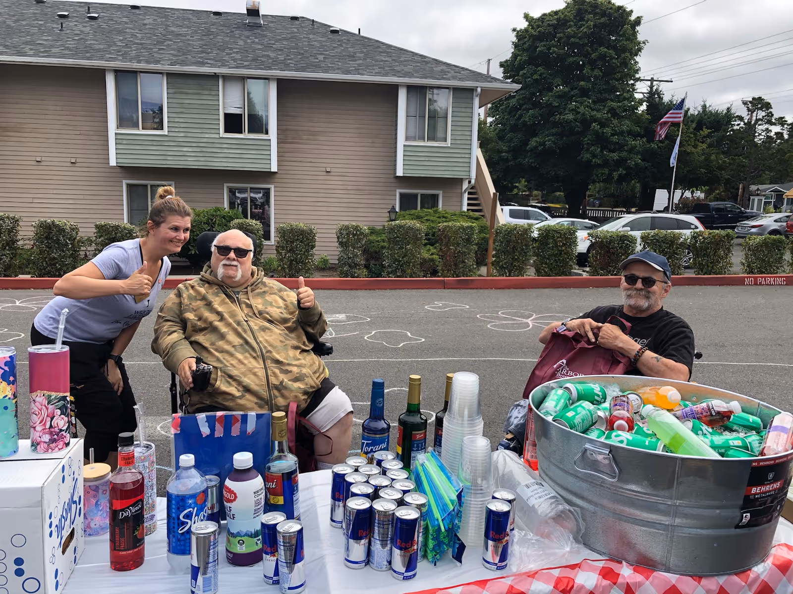 Two elderly men sitting outdoors at a table covered with a red and white checkered tablecloth, displaying various beverages including cans of Red Bull, bottles of syrup, and a large metal tub filled with assorted canned and bottled drinks. A woman stands next to one of the men, smiling and giving a thumbs-up. Behind them is a residential building, bushes, parked cars, and trees under a cloudy sky.