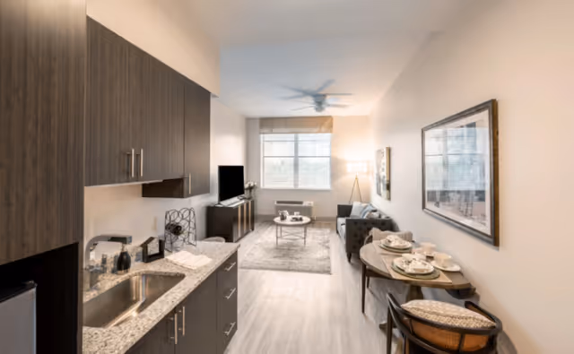 A modern studio apartment with a kitchenette on the left featuring dark wood cabinets, a granite countertop, and a stainless steel sink. The living area includes a gray sofa, a coffee table on a rug, a TV on a stand, and a large window with blinds. On the right side, there is a small dining table set for two with plates, cups, and napkins. The walls are light-colored with framed artwork, and a ceiling fan is visible above.