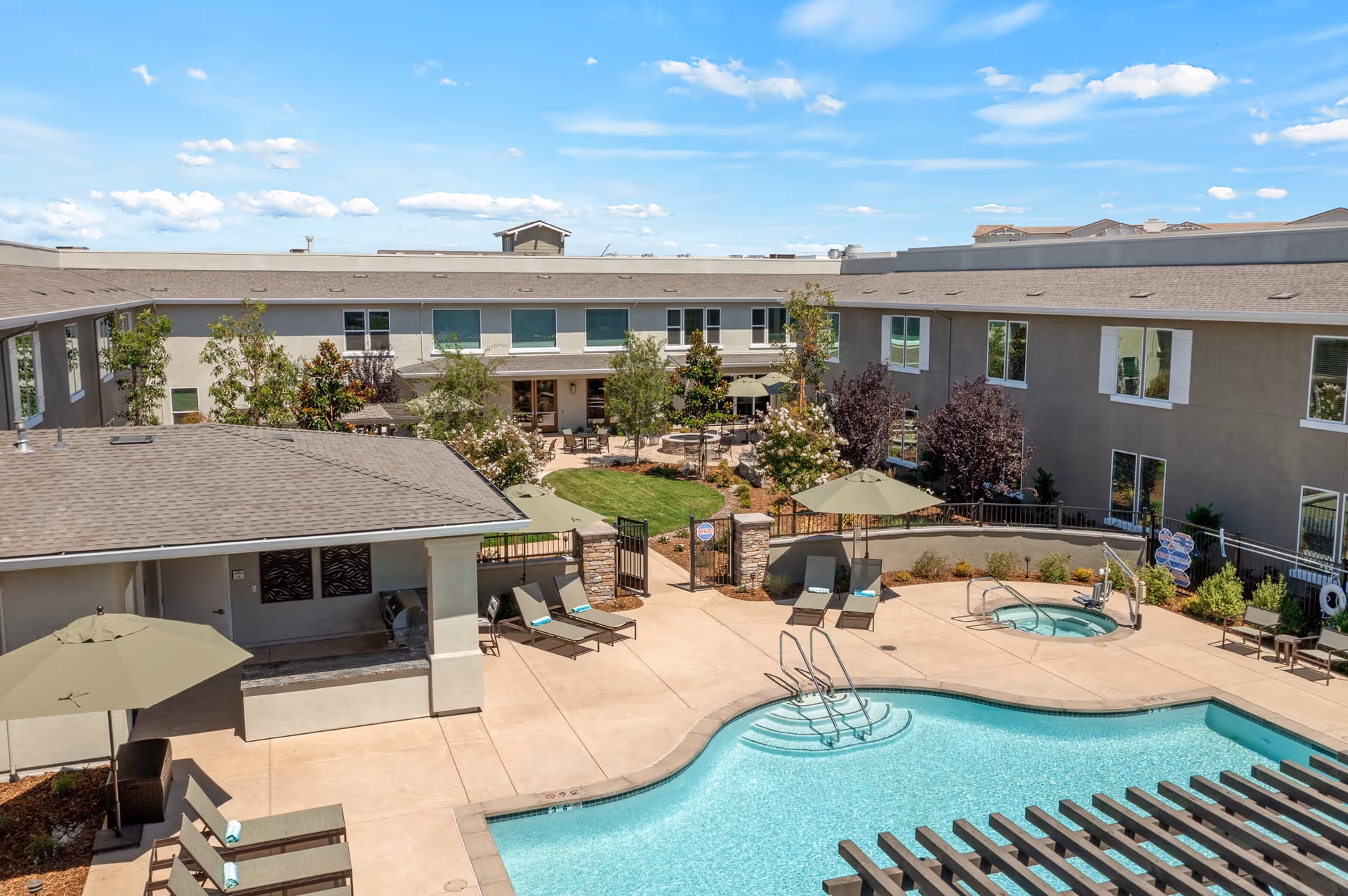 Outdoor courtyard area of Oakmont of Lodi featuring a swimming pool, hot tub, lounge chairs with umbrellas, a shaded seating area, and surrounding two-story building with windows. The sky is clear with some clouds.