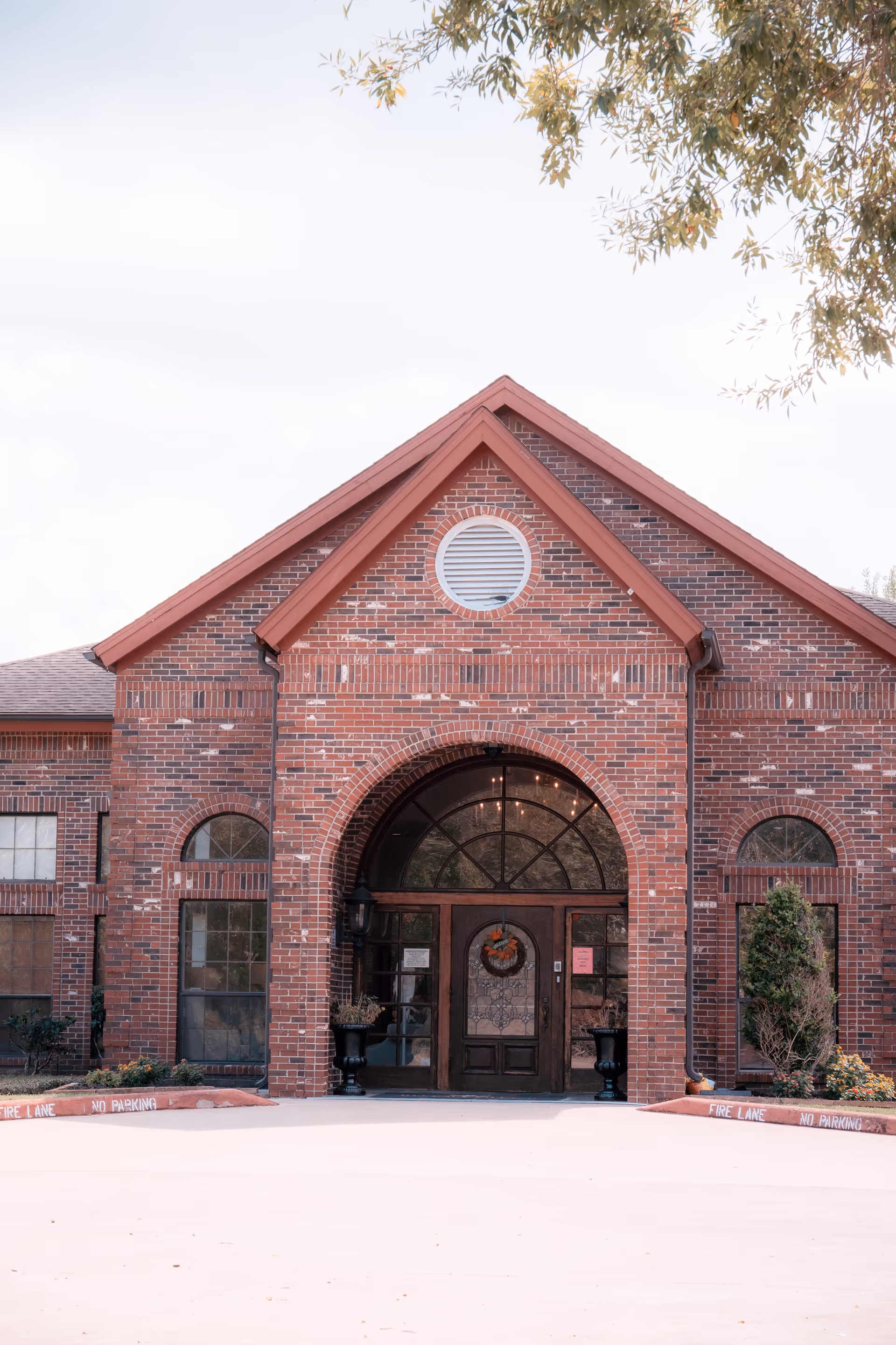Front exterior view of a brick building with a large arched entrance door decorated with a wreath. The building has multiple windows and a triangular roof section with a circular vent. There are small plants and bushes on either side of the entrance and a concrete driveway in front marked with 'FIRE LANE NO PARKING'.