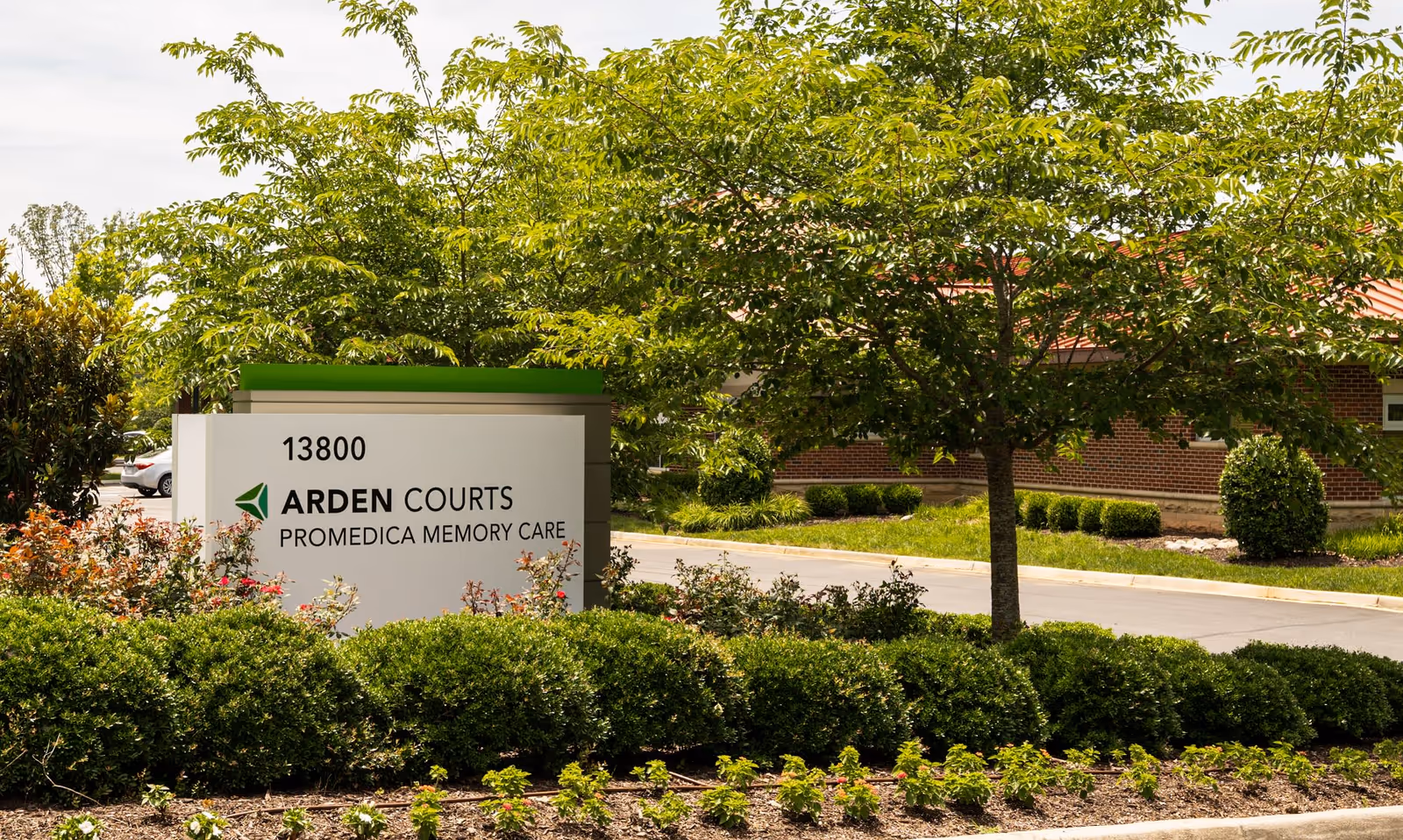 Outdoor view of Arden Courts Promedica Memory Care facility sign surrounded by green bushes and trees, with part of the building and a parking lot visible in the background.