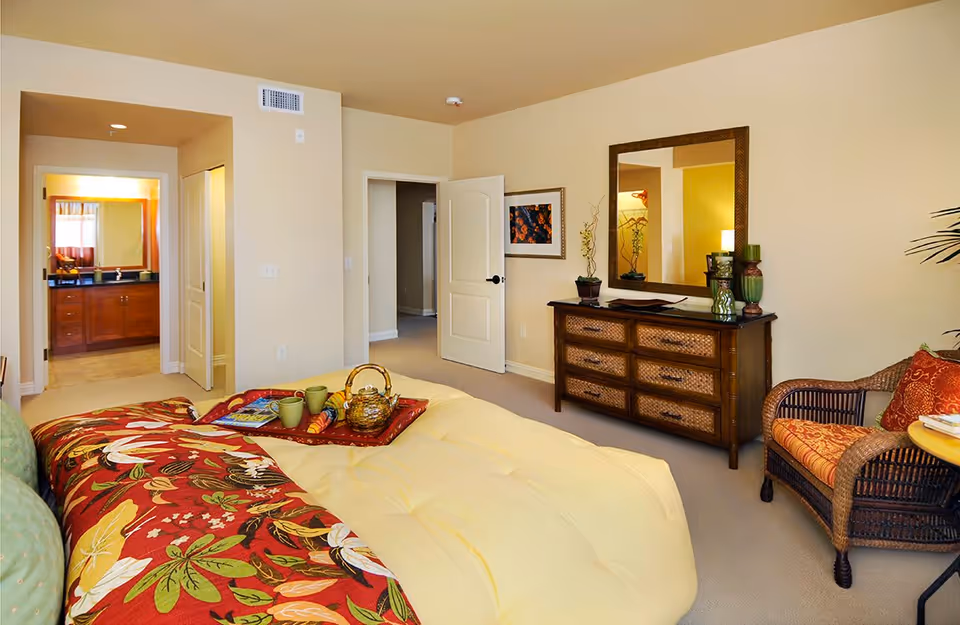 A cozy bedroom in a retirement community featuring a large bed with a yellow comforter and a colorful floral blanket. The room includes a wooden dresser with a large mirror above it, a wicker armchair with orange cushions, and a small round table. The bedroom opens into a bathroom with a wooden vanity and a mirror, visible through an open doorway.