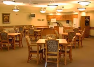 A dining room with multiple wooden tables and cushioned chairs arranged neatly. The room has warm lighting with ceiling fixtures, a kitchen area with cabinets, a microwave, and a stove in the background. The walls are decorated with framed artwork and small decorative items.