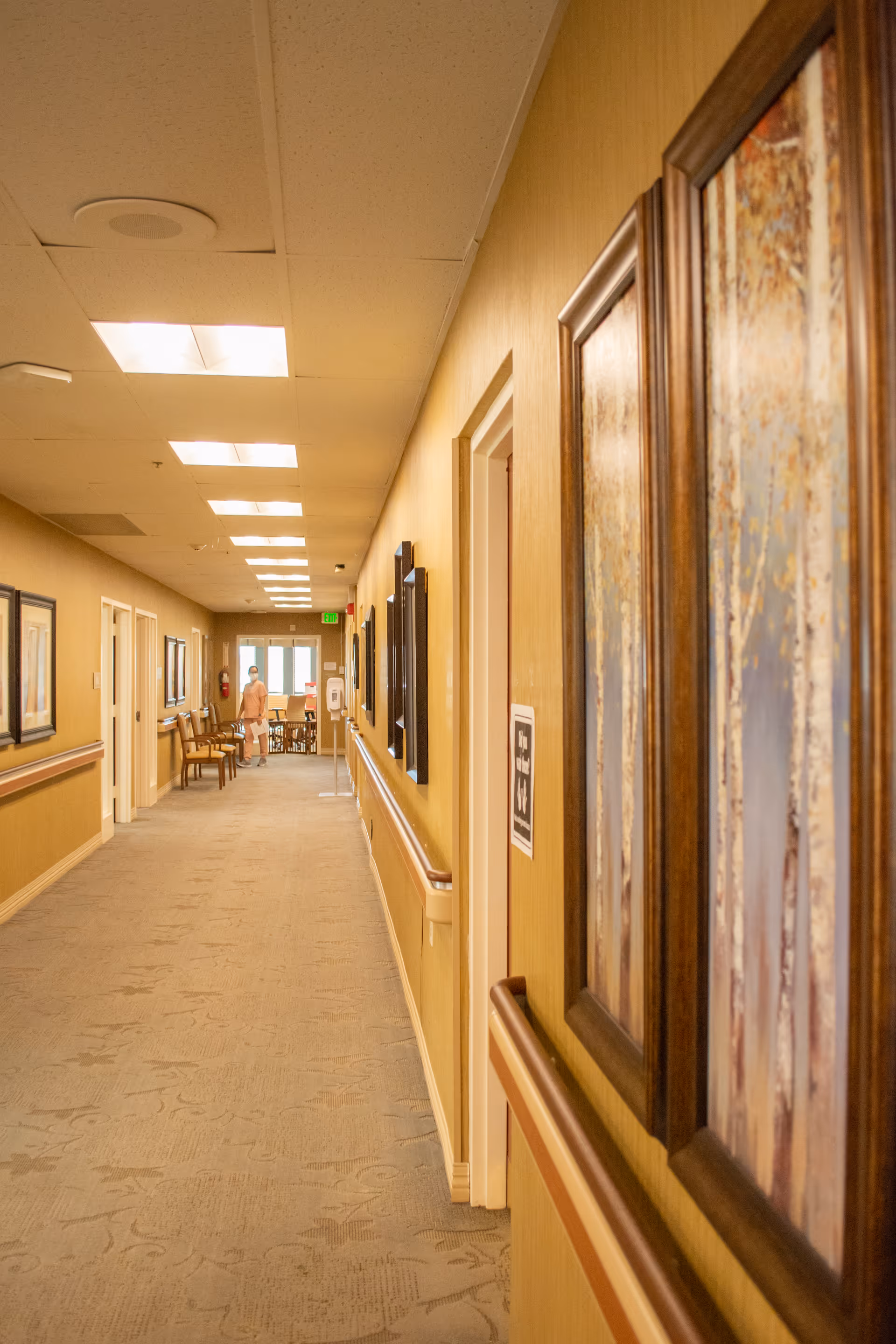 Long, well-lit interior hallway of a care facility with handrails, framed artwork, chairs and a person visible at the far end.