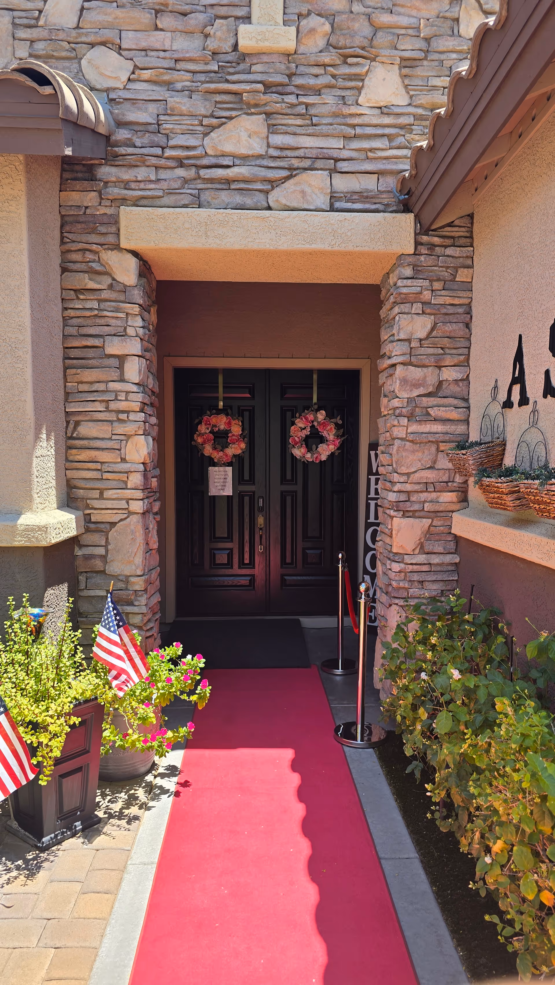 Stone-faced entryway with double dark doors decorated with floral wreaths, a red carpet runner and potted plants with small American flags.