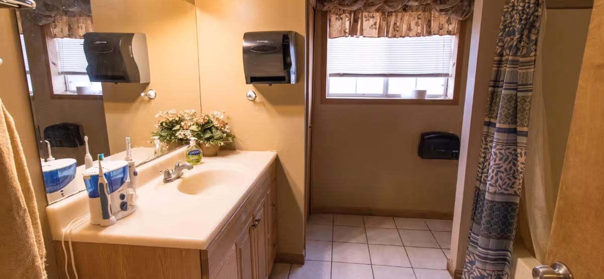 A bathroom with a single sink vanity featuring a white countertop and wooden cabinets. On the countertop are two electric toothbrushes and a container of hand soap. Above the sink is a large mirror with a paper towel dispenser mounted on the wall. A window with blinds and a floral valance is visible, along with a shower area with a patterned curtain. The floor is tiled, and there is a towel hanging on the left side.