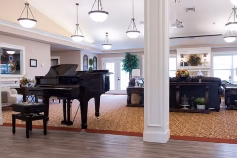Spacious assisted living facility common area with a black grand piano and matching bench on a wooden floor. The room features patterned carpet, multiple hanging ceiling lights, a white column, comfortable seating including sofas, a fireplace with floral decorations, and large windows letting in natural light.