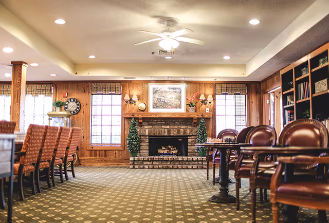 A cozy living room area with a brick fireplace in the center, flanked by two tall green plants. The room has wood-paneled walls, two windows with blinds, and a ceiling fan with lights. On the left side, there is a long table with several patterned chairs, and on the right side, there are leather chairs and a bookshelf filled with books and decorative items.
