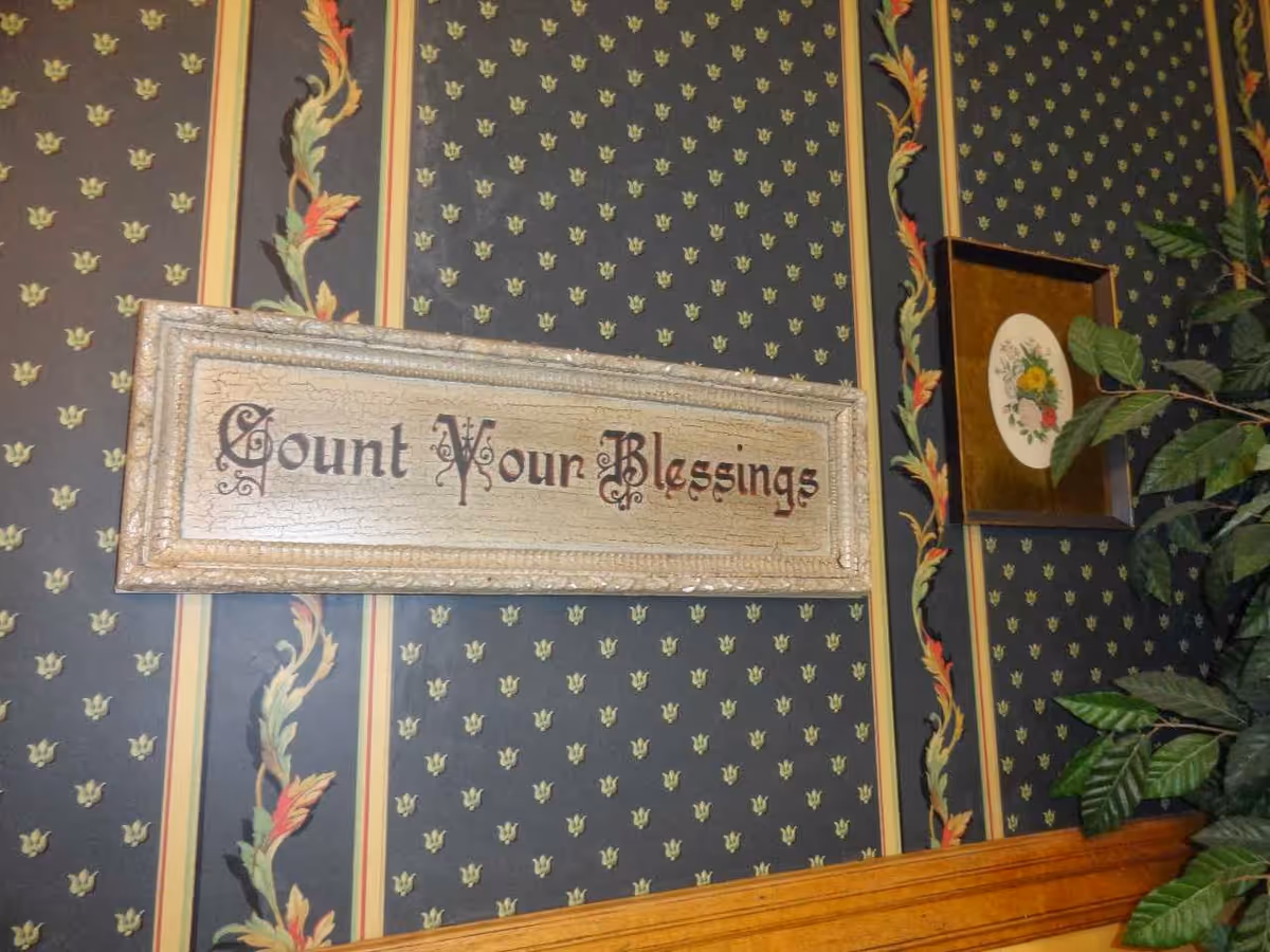 A decorative wall with patterned wallpaper featuring small floral motifs and vertical stripes with leaves. A framed sign on the wall reads 'Count Your Blessings' in ornate lettering. To the right, there is a framed floral artwork and part of a green leafy plant is visible.
