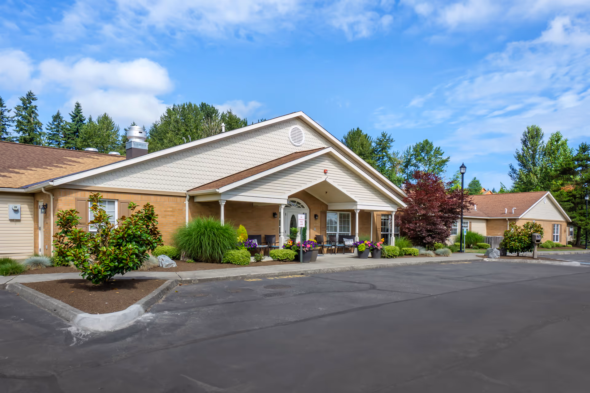 Exterior view of a single-story senior living facility building with a covered entrance, outdoor seating, and landscaped plants and flowers under a partly cloudy blue sky.