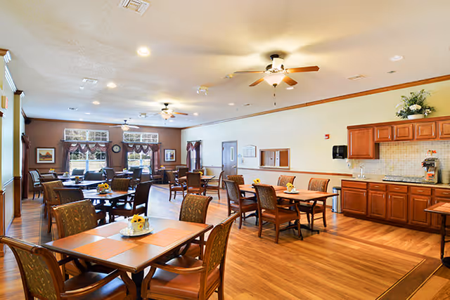 A spacious dining room with multiple wooden tables and chairs arranged neatly. The room has hardwood flooring, ceiling fans with lights, and large windows with decorative curtains allowing natural light to enter. There is a kitchenette area with wooden cabinets, a countertop, and a coffee machine on the right side. The walls are painted in light colors with some framed artwork and floral decorations.