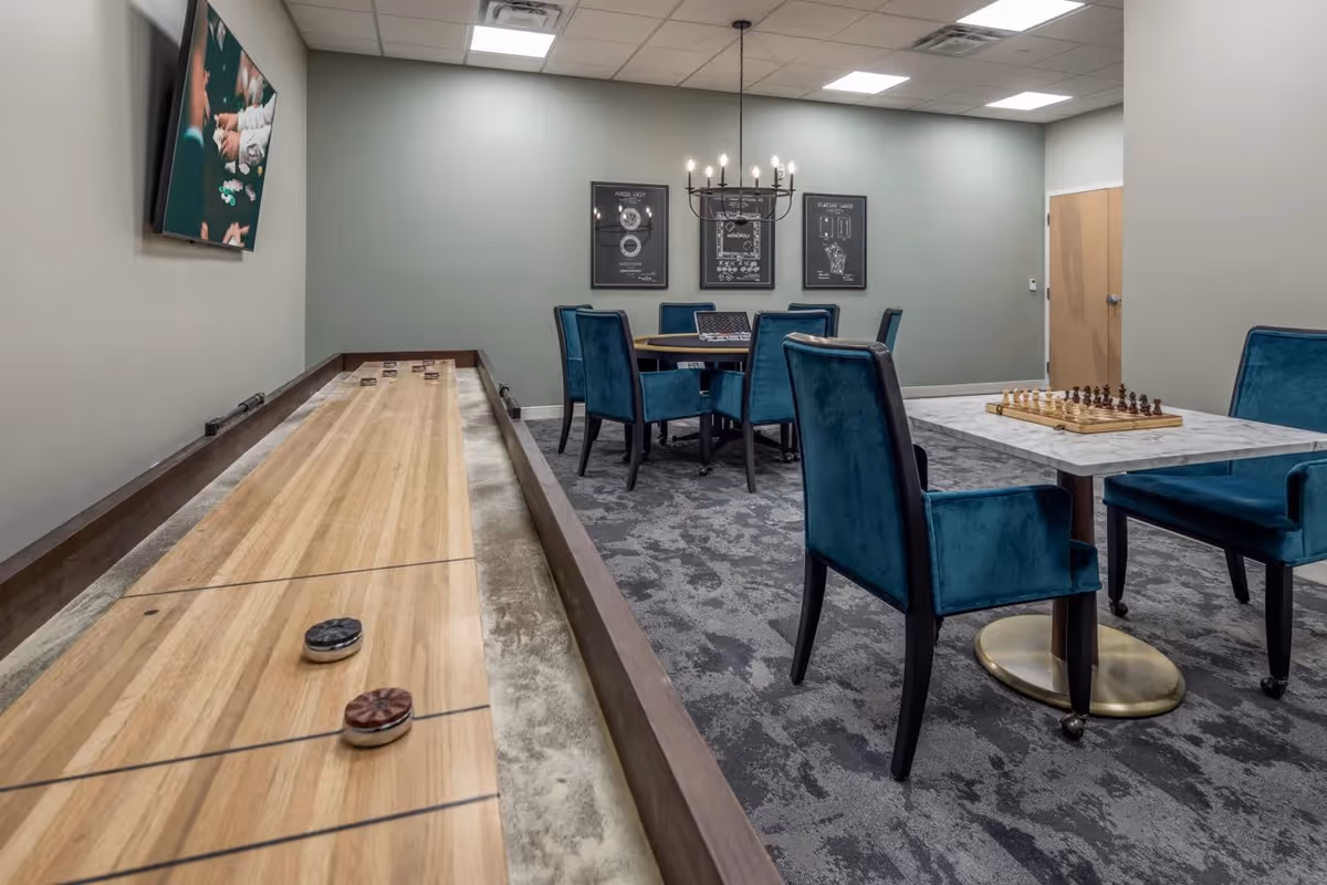 A game room with a shuffleboard table in the foreground and a marble table with a chess set on it. There are several blue upholstered chairs around the tables, a round table with chairs in the background, and framed artwork on the wall. The room has a carpeted floor and ceiling lights.