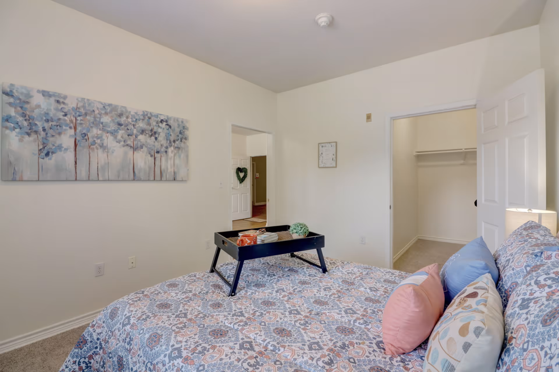 A bedroom with a bed covered in a patterned blue and white bedspread with several pillows. A black tray with a cup, books, and a small plant is on the bed. The room has beige walls, a painting of trees on one wall, an open door leading to a closet, and another open doorway showing a hallway with a white door decorated with a green heart-shaped wreath.