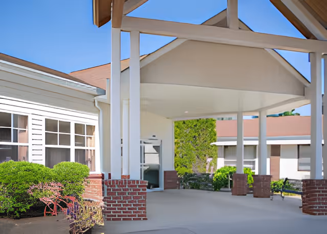 Covered entrance/porte-cochère of a senior living facility with white columns, brick bases, and surrounding shrubs.