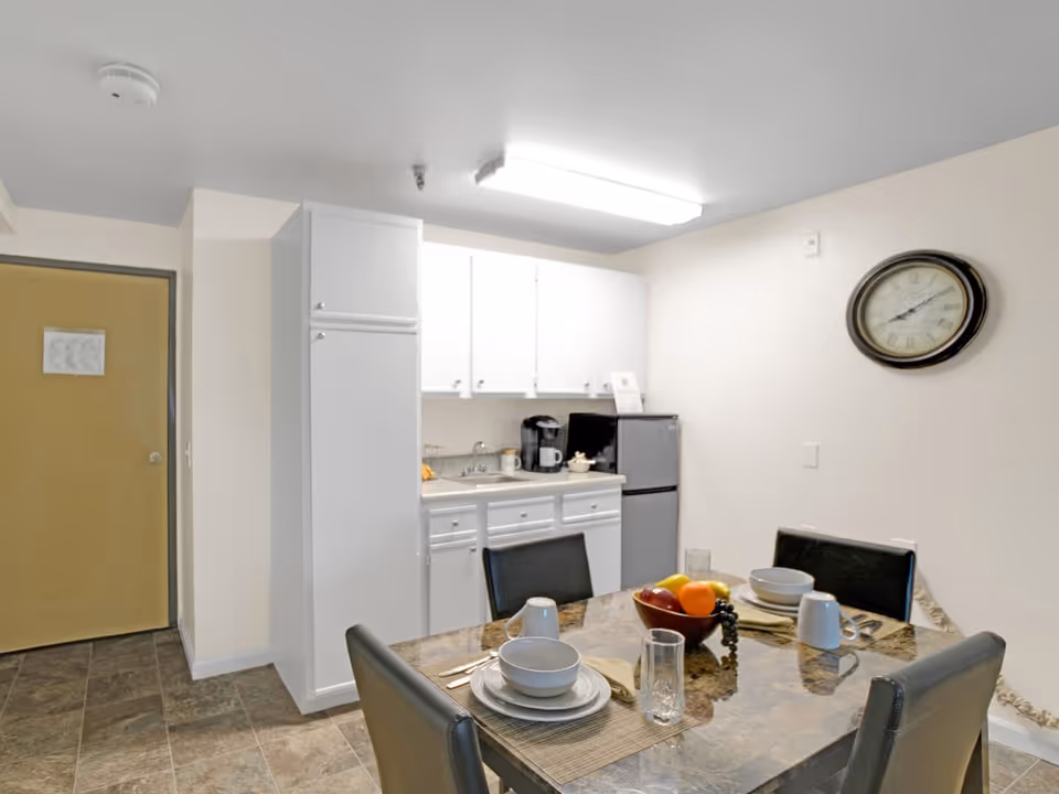 A small kitchen and dining area with white cabinets, a refrigerator, a coffee maker, and a sink. In the foreground, there is a dining table set with plates, bowls, cups, glasses, and a bowl of fruit. A round wall clock is mounted on the wall above the table.