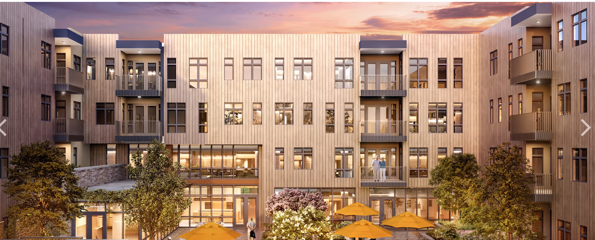 Exterior view of a modern senior living facility at sunset with multiple balconies, large windows, trees, and yellow patio umbrellas in the courtyard.