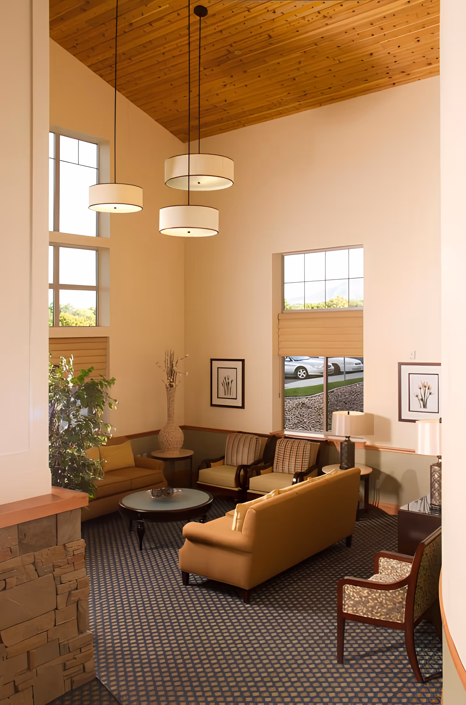 A cozy living room area with a high wooden ceiling and three hanging round pendant lights. The room features a tan sofa, two cushioned armchairs, a patterned armchair, a round glass coffee table, and side tables with lamps. There are framed botanical prints on the walls and large windows letting in natural light, with a view of parked cars outside. A tall decorative vase and a green plant add to the decor.