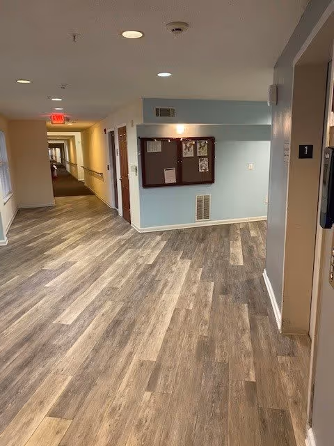 Interior hallway of a senior living facility with wood-patterned flooring, light blue and beige walls, ceiling lights, a bulletin board on the wall, and doors along the corridor.