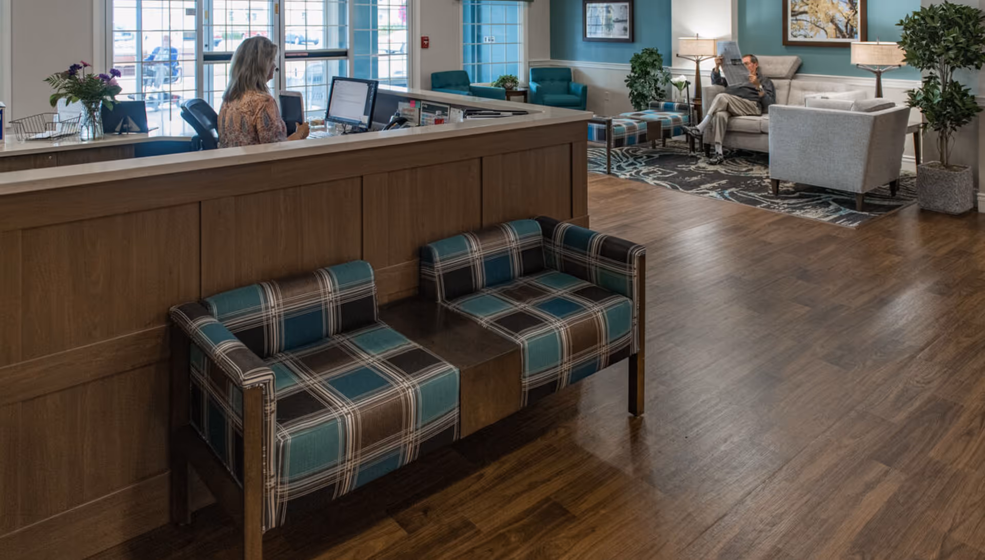Reception desk and adjacent lounge in a senior living facility with a plaid bench, sofa seating, wood floors, and staff at the desk.