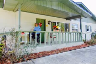 A person sitting on a chair on the covered front porch of a single-story building with green railings and a green door. There are potted flowers on the porch and some plants in front of the porch. The building has white exterior walls and a sloped roof.