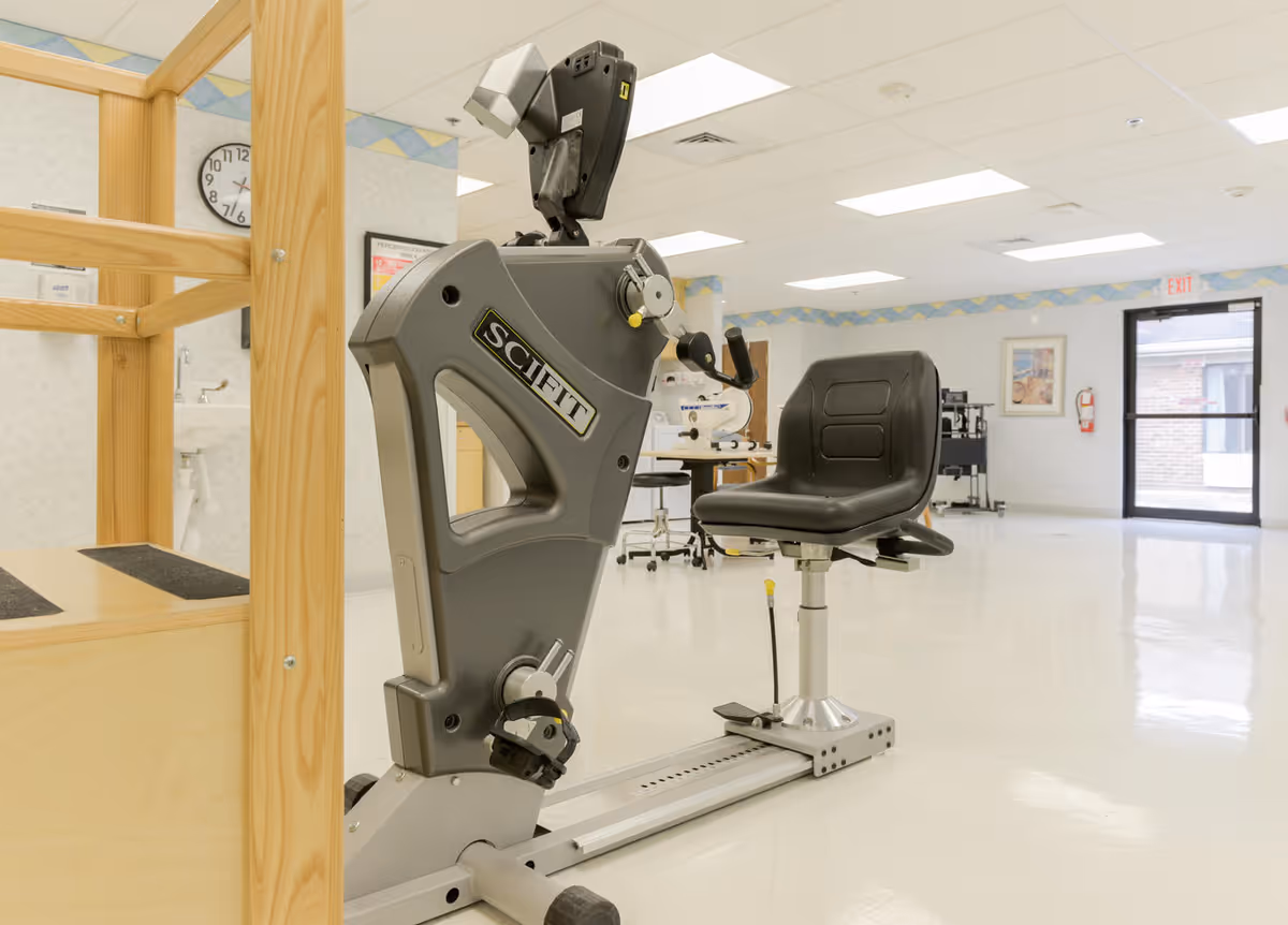 A physical therapy or rehabilitation room with a SciFit exercise machine featuring a black seat and pedals. The room has a clean, bright interior with white floors, light-colored walls, a clock on the wall, and an exit door leading outside.