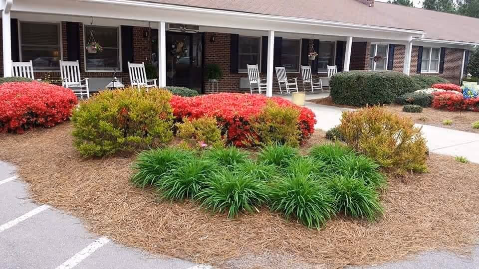 Front entrance of a single-story assisted living building with a covered porch, rocking chairs, and landscaped shrubs and flowers.