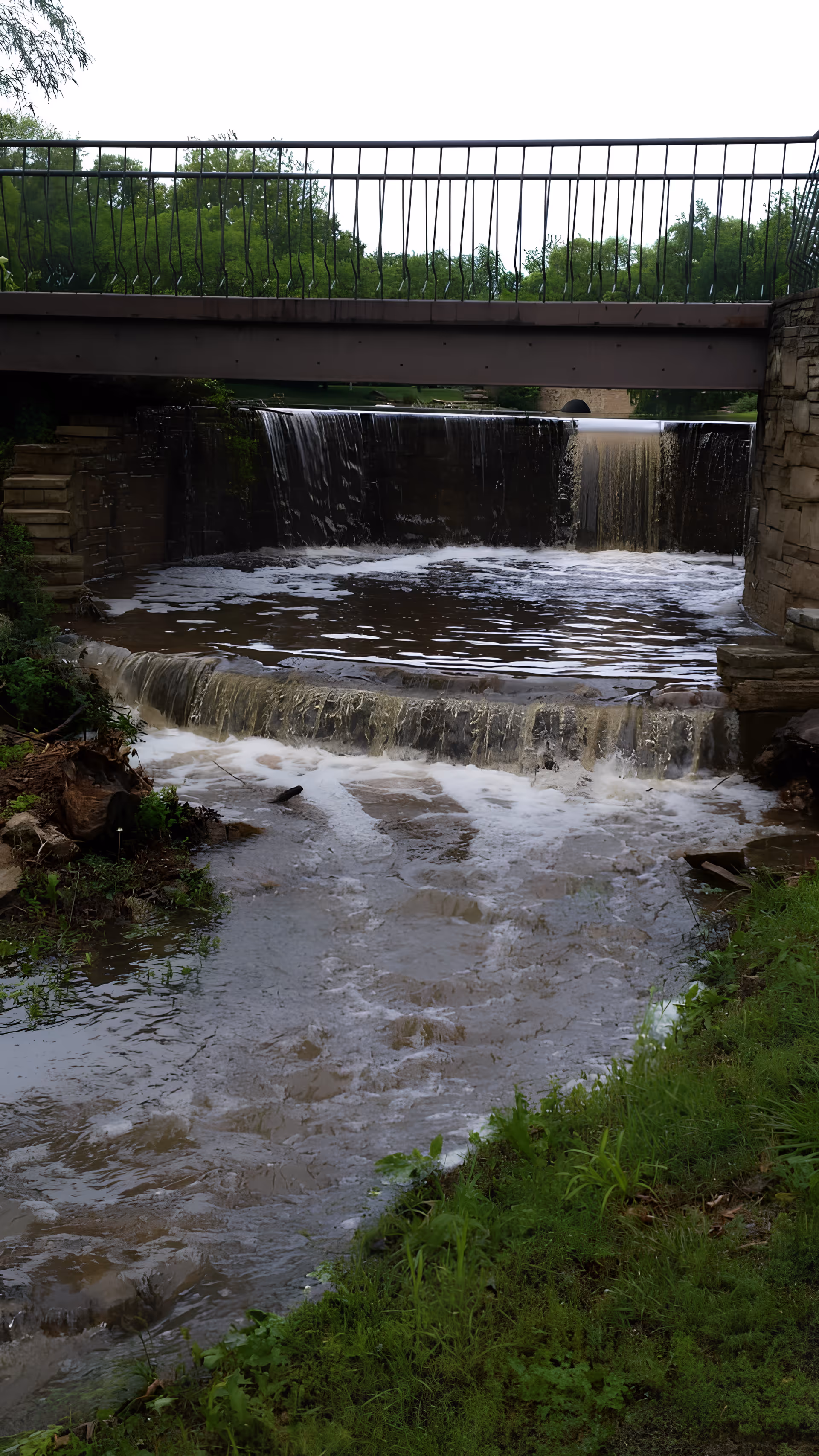 A small waterfall under a metal bridge with stone walls on either side, surrounded by green grass and trees in the background.